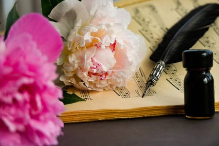 close up of piano sheet music with vintage quill pen resting on it and spare black ink surrounded by fragrant fresh peonies creating a creative mood.