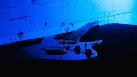 Schwarze Hochglanzgitarre wartet auf dem Tisch mit blauem Hintergrund. Die Gitarre ruht und ist bereit für das Rockkonzert. Akustische Gitarrenkomposition mit blauem und schwarzem Hintergrund.