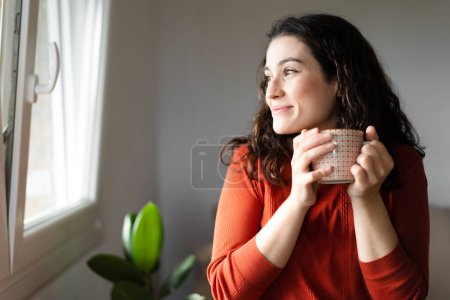 Foto de Joven feliz hermosa mujer sosteniendo una taza de bebida caliente y mirando a través de la ventana se siente bien - Imagen libre de derechos