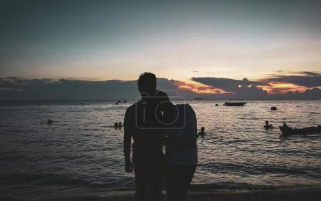 Foto de Una fotografía de una pareja de pie en la playa al atardecer, hay dos personas de pie en la playa viendo la puesta de sol. - Imagen libre de derechos