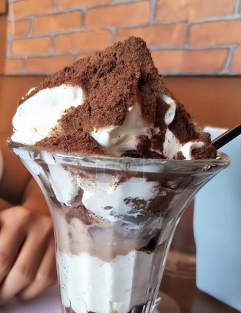 Téléchargez les photos : Une photographie d'un sundae au dessert avec crème fouettée et chocolat, un peu de dessert dans une tasse en verre avec une cuillère sur une table. - en image libre de droit
