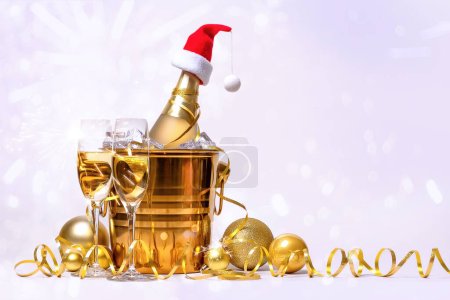 A bottle of champagne with a red santa claus cap in a golden bucket with ice and two glasses on a white background.