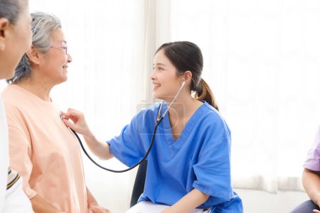 Téléchargez les photos : Infirmière asiatique utilisant un stéthoscope pour vérifier la fréquence cardiaque de la patiente âgée à la maison. Assistance médicale. Concept de maison de soins infirmiers pour aidants naturels. - en image libre de droit