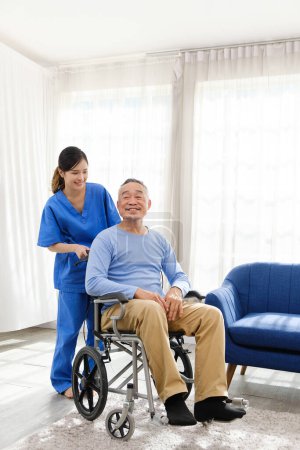 Photo for The caregiver therapist stands with an Asian senior sitting in a wheelchair with an empty sofa in the background. The nursing home facilitates a support group. - Royalty Free Image