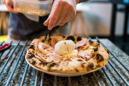 Neapolitan pizza style: close-up chef hand seasoning crushed pistachio over mortadella and burrata pizza on the pizza cooling rack. Focus on burrata and mortadella. Italian baked Napoli pizza.