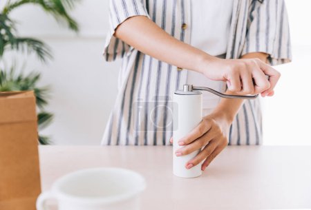 Téléchargez les photos : Recadrer la vidéo d'une femme utilisant des moulins à café de couleur blanche sur la table avant de verser sur le café moulu sur le filtre à papier sur un goutteur de verre. Une méthode alternative est appelée Dripping. - en image libre de droit