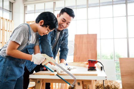 Asian father and son work as a woodworker or carpenter, Father teaches his son to saw a wooden plank with hack saw carefully together with teamwork. Craftsman carpentry working at home workshop studio