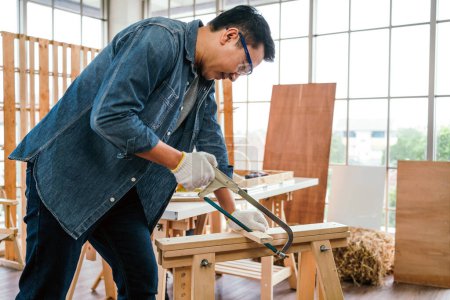 Asian father and son work as a woodworker or carpenter, Father wears safety goggles and saw a wooden plank with hacksaw carefully. Craftsman carpentry working at home workshop studio. Small business