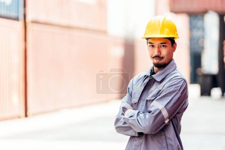 Atractivo ingeniero industrial japonés en sombrero amarillo duro, uniforme de seguridad con un patio de contenedores borroso en el fondo. Trabajando en el centro logístico. Inspector, supervisor en terminal de contenedores