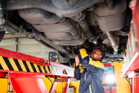 Portrait of an African female mechanic in yellow and blue uniform standing under the car bottom for inspecting in garage. Woman smiling while holding a wrench. Car repair service. Vehicle maintenance