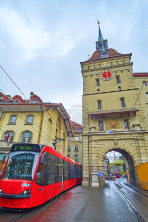 Foto de BERN, SUIZA - 31 DE MARZO DE 2022: Paseos modernos en tranvía rojo en Waisenhausplatz junto a la torre medieval de Kafigturm, el 31 de marzo en Berna, Suiza - Imagen libre de derechos