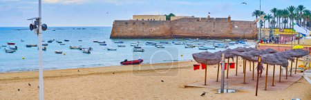 Téléchargez les photos : Panorama de la plage de sable La Caleta avec de petits bateaux de pêche au bord de la baie de Cadix et le château de Santa Catalina en arrière-plan, Espagne - en image libre de droit