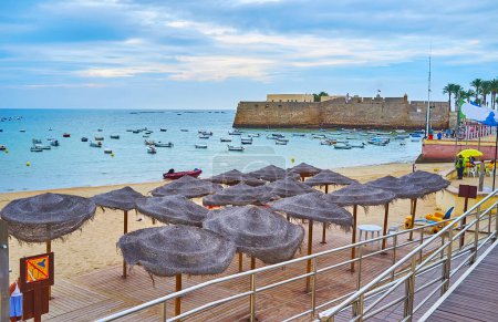 Téléchargez les photos : Les parasols en paille sur la plage de La Caleta, les petits bateaux de pêche, balançant sur les vagues et le château de Santa Catalina en arrière-plan, Cadix, Costa de la Luz, Espagne - en image libre de droit