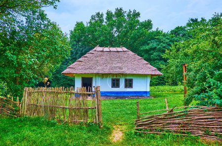 Foto de La antigua cerca de mimbre, pequeño patio verde y la casa de hata pueblo encalado con techo de paja y árboles exuberantes en el fondo, Chernivtsi scansen, Ucrania - Imagen libre de derechos