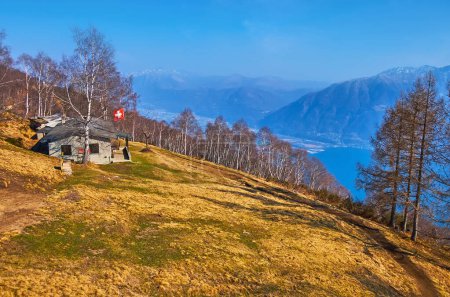 El seco prado montano amarillo de principios de primavera en la ladera del monte Cimetta con la pequeña casa contra el abedul y el lago Maggiore, Locarno, Suiza