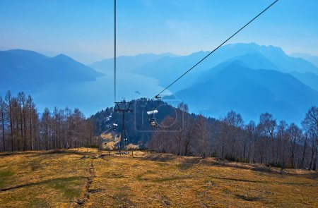 The sunny and hazy mountain landscape and Lake Maggiore from the Cimetta Mount chairlift, riding along the dry yellow montane meadow, Ticino, Switzerland