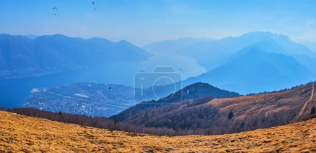 Panorama vom Gipfel des Monte Cimetta mit ausgetrockneter Bergwiese am Berghang und nebelblauem Lago Maggiore im Tal, Tessin, Schweiz