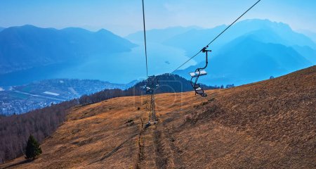 Die Berglandschaft und der nebelblaue Lago Maggiore vom Sessellift Cimetta Mount, Fahrt entlang der trockenen gelben Bergwiese, Tessin, Schweiz