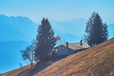 Das kleine Chalet-Gästehaus am Hang von Cardada Cimetta vor den hellblauen, dunstigen Alpen, Tessin, Schweiz