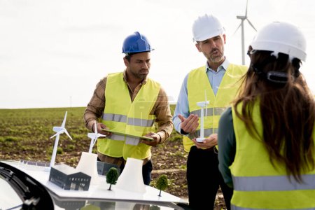 Foto de Tres ingenieros caucásicos y latinos de pie en el campo de la turbina eólica y discutiendo animados sobre modelos de plástico - Imagen libre de derechos