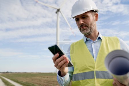 Téléchargez les photos : Ingénieur masculin caucasien debout sur le champ des éoliennes et utilisant un téléphone mobile - en image libre de droit
