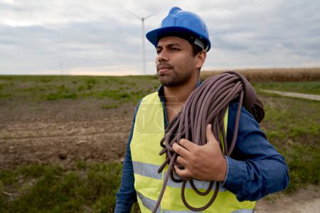 Téléchargez les photos : Technicien d'entretien latin portant une corde d'escalade et marchant à travers l'éolienne - en image libre de droit