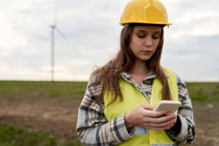 Téléchargez les photos : Ingénieur femme caucasienne debout sur le champ d'éoliennes et utilisant le téléphone mobile - en image libre de droit