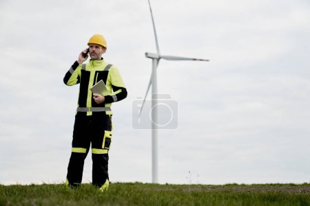 Téléchargez les photos : Ingénieur caucasien debout sur le champ de l'éolienne, tenant tablette numérique et passer un appel - en image libre de droit