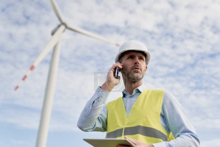 Téléchargez les photos : Ingénieur masculin caucasien debout sur le champ des éoliennes avec tablette numérique et parler au téléphone - en image libre de droit