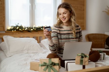 Téléchargez les photos : Femme caucasienne assise sur le lit et faisant des achats de Noël avec l'utilisation d'un ordinateur portable et carte de crédit - en image libre de droit