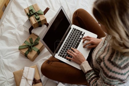 Draufsicht einer kaukasischen Frau, die auf dem Bett sitzt und zwischen Weihnachtsgeschenken im Laptop blättert