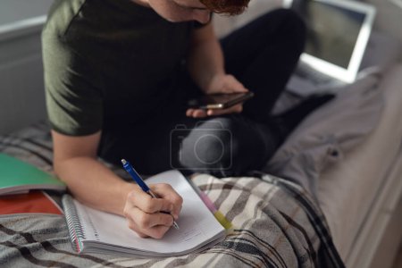 Téléchargez les photos : Adolescent caucasien garçon assis sur le lit et apprendre à l'aide de téléphone - en image libre de droit