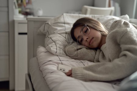 Photo for Sad caucasian teenage girl lying on bed and looking at her phone held in hand - Royalty Free Image