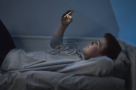 Téléchargez les photos : Joyeux caucasien adolescent garçon regarder téléphone mobile tandis que couché dans son lit la nuit dans sa chambre - en image libre de droit