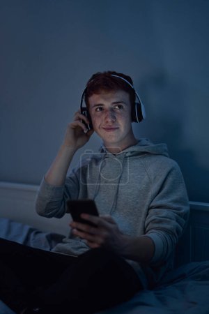 Foto de Caucásico adolescente usando auriculares y escuchando música en la noche en el dormitorio - Imagen libre de derechos