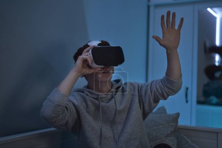 Téléchargez les photos : Adolescent caucasien garçon en utilisant virtuel réalité lunettes tandis que assis dans chambre à coucher la nuit - en image libre de droit