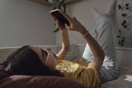 Téléchargez les photos : Caucasien adolescent fille navigation téléphone tout allongé sur le lit avec ses jambes vers le haut - en image libre de droit