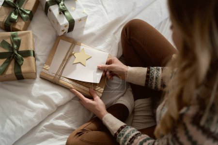 Hochwinkelaufnahme einer kaukasischen Frau, die auf dem Bett sitzt und Weihnachtsgeschenk mit Grußkarte verpackt 