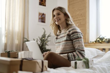 Téléchargez les photos : Vue à angle bas de la femme caucasienne assise sur le lit et naviguant ordinateur portable parmi les cadeaux de Noël - en image libre de droit