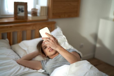 Téléchargez les photos : Femme caucasienne utilisant le téléphone mobile et couché au lit le matin - en image libre de droit