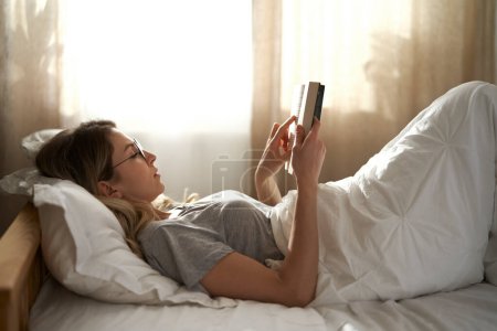 Photo for Caucasian woman lying down in bed and reading book at the morning - Royalty Free Image