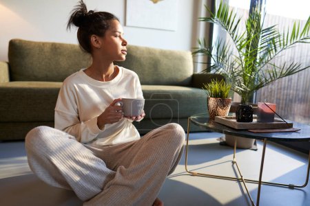 Téléchargez les photos : Mixed race woman sitting at the floor at home and enjoying the coffee - en image libre de droit