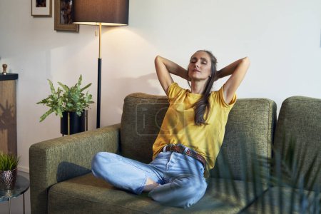 Téléchargez les photos : Jeune femme assise et relaxante sur un canapé avec les mains derrière la tête et les yeux fermés - en image libre de droit