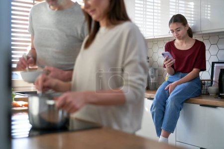 Photo for Teenage daughter using mobile phone in kitchen when parents cooking together - Royalty Free Image
