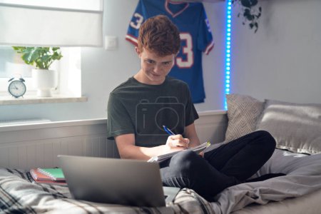 Téléchargez les photos : Adolescent caucasien garçon assis sur le lit et apprendre à l'aide d'un ordinateur portable - en image libre de droit