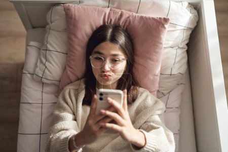Téléchargez les photos : Top vue de caucasien adolescent fille prendre selfie avec téléphone tandis que couché sur le lit sur son dos - en image libre de droit