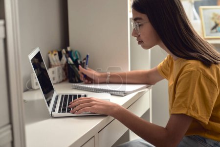 Téléchargez les photos : Caucasienne adolescente assis sur le bureau dans la chambre et tapant sur ordinateur portable - en image libre de droit