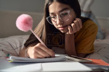 Photo for Caucasian teenage girl lying on front on bed and making notes - Royalty Free Image