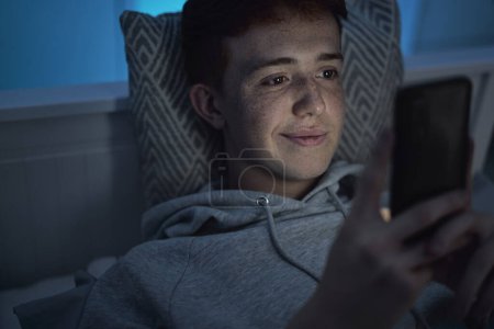 Téléchargez les photos : Joyeux caucasien adolescent garçon à l'aide de téléphone mobile tout couché dans son lit la nuit dans sa chambre - en image libre de droit