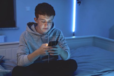 Photo for Affectionate caucasian teenage boy using mobile phone while sitting at night in his room - Royalty Free Image